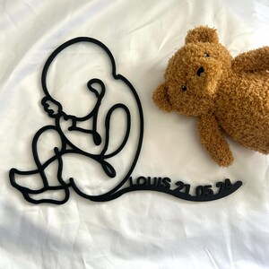 Puede incluir: Una escultura de arte lineal minimalista negra de un feto, con el nombre "LOUIS 21.05.24" recortado debajo. Un oso de peluche marrón está acostado junto a la escultura sobre una superficie blanca. La escultura es un objeto decorativo.