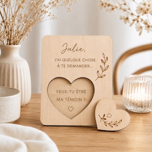 Peut inclure: Une plaque rectangulaire en bois clair gravée avec le texte "Julie, J'AI QUELQUE CHOSE À TE DEMANDER..." et "VEUX-TU ÊTRE MA TÉMOIN ?", comprenant une découpe en forme de cœur et un petit cœur en bois assorti orné d'un motif de feuilles.