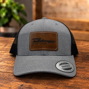 May include: A heather gray baseball cap with a black mesh back and a brown rectangular leather patch on the front. The patch features the black text "Dillinger SYSTEMS". A circular black and silver sticker on the brim reads "THE AUTHENTIC YUPOONG SNAPBACK".