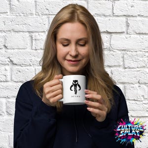 May include: A white ceramic mug featuring a black Mythosaur skull graphic and text printed on the front. The mug has a cylindrical body with a handle on the side, displaying a clean design against the white surface of the drinkware.