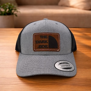 May include: A heather gray snapback cap with black mesh back panels. The front features a rectangular brown leather patch with the black text THE DARK SIDE and a partial circular graphic. A silver and black sticker is on the brim.