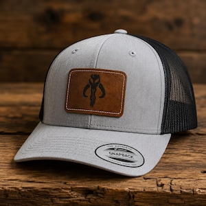 May include: A gray and black trucker hat featuring a rectangular brown leather patch on the front with a black Mythosaur skull emblem. The brim has a circular sticker that reads The Authentic Yupoong Snapback Since 1974.