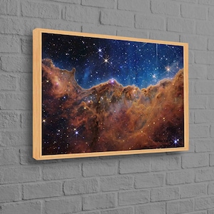 May include: A framed canvas print featuring a vibrant depiction of a nebula in space. The artwork showcases a blend of orange, brown, and blue hues, with numerous stars scattered across the dark background. The print is encased in a light-colored wooden frame.