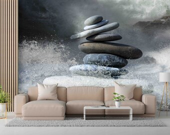 May include: A stack of gray stones balanced on top of each other in a zen garden setting. The stones are surrounded by water and a white surface. The background is a blurry image of a waterfall.