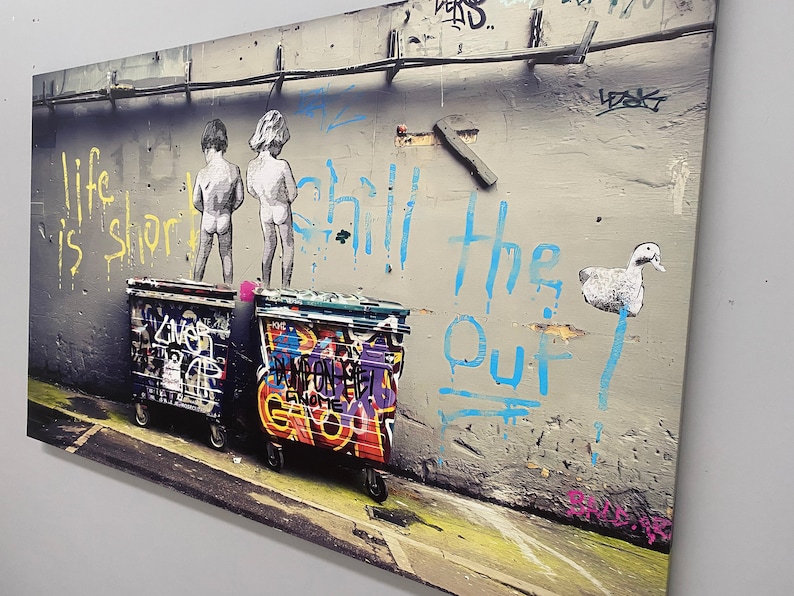 May include: A street art image with the text "Life is short, chill the f out" in blue paint on a gray wall. Two figures are painted on the wall, and there is a white duck painted on the wall. Two graffiti-covered trash cans are in front of the wall.
