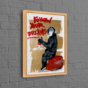 Könnte beinhalten: Ein gerahmter Druck eines Schimpansen, der auf einer roten Coca-Cola-Kiste sitzt und eine Sprühdose hält. Der Schimpanse trägt ein schwarzes Hemd und hat einen goldenen Hintergrund. Der Text "Follow Your Dreams" ist in roter Sprühfarbe auf den Hintergrund geschrieben.