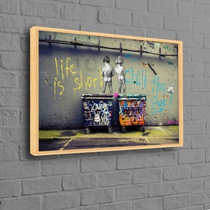 May include: A framed print of a street art piece featuring two children standing in front of two graffiti-covered trash cans. The wall behind them has the words "Life is short, chill the f*** out" written in yellow and blue paint.