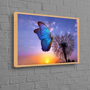May include: A framed print depicting a blue butterfly with detailed wings on a dandelion clock. The backdrop shows a sunset with orange and purple tones, set against a blue sky, all within a wooden frame.