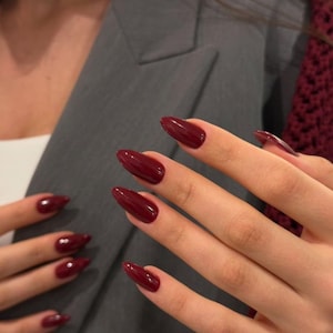 May include: Close-up of hands with long, almond-shaped nails painted a deep burgundy color. The nails are glossy and reflect light. The hands are positioned in front of a gray blazer.