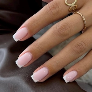 Op de afbeelding: Close-up van een hand met French manicure. De nagels zijn zachtroze met witte punten. Twee gouden ringen sieren de vingers. De achtergrond is een donkerbruin leren oppervlak.