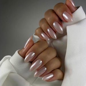 Puede incluir: Manos con uñas largas en forma de almendra, pintadas de rosa nacarado. Las uñas tienen un acabado brillante. El fondo es blanco.