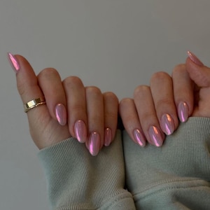 Puede incluir: Manos con uñas rosas iridiscentes de forma ovalada. Un anillo dorado está en el dedo índice de la mano izquierda. Las uñas tienen un acabado brillante, que refleja la luz. Las manos están sobre un fondo verde claro.