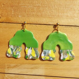 Könnte beinhalten: Ein Paar grüne und weiße abstrakte Wirbelohrringe mit goldenen Haken. Die Ohrringe sind bogenförmig.