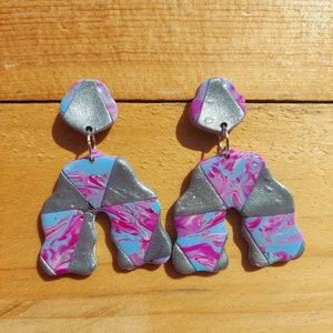 Könnte beinhalten: Ein Paar handgefertigte Polymer-Ton-Ohrringe. Jeder Ohrring hat ein geometrisches Design mit einem silbernen Oberteil und einem marmorierten Muster aus Pink, Blau und Silber. Die Ohrringe sind an silberfarbenen Haken aufgehängt.