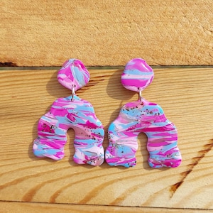 Könnte beinhalten: Ein Paar abstrakte, schwingende Ohrringe in Pink, Blau und Silber. Die Ohrringe sind aus Polymer Clay gefertigt und haben eine einzigartige, organische Form.