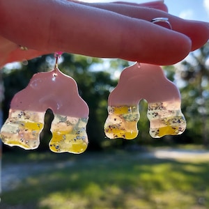 Könnte beinhalten: Ein Paar rosa Harzohrringe mit einem wellenförmigen, bogenförmigen Design. Die Ohrringe haben eine Schicht aus klarem Harz mit eingebetteten goldenen und silbernen Glitzer.