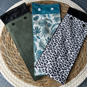 May include: Three fabric cases with snap closures. One is olive green corduroy, another features a tropical leaf print on a white background, and the third has a black and white leopard print. All are displayed on a woven placemat.