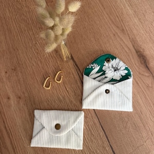 Può includere: Due piccole bustine in velluto a coste bianco a forma di busta con chiusure a scatto in ottone, una chiusa e una aperta che rivela una fodera in tessuto floreale verde e bianco, accompagnate da un paio di orecchini a cerchio dorati e un mazzetto di erba secca.