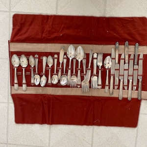 May include: A set of silver flatware in a red and beige roll-up case. The set includes spoons, forks, and knives.