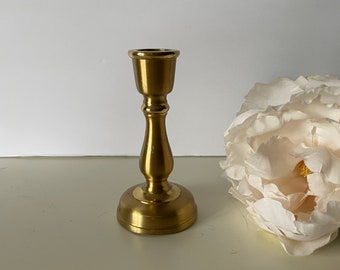 May include: A gold metal candle holder with a simple design. The holder has a round base and a tall, slender stem.