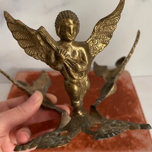 May include: A gold-toned metal candle holder with three angel figures. Each angel has wings and is holding a candle. The candle holder is on a red velvet surface.