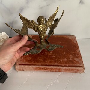 May include: A gold-toned metal angel figurine with wings outstretched. The angel is holding a musical instrument and is standing on a decorative base.