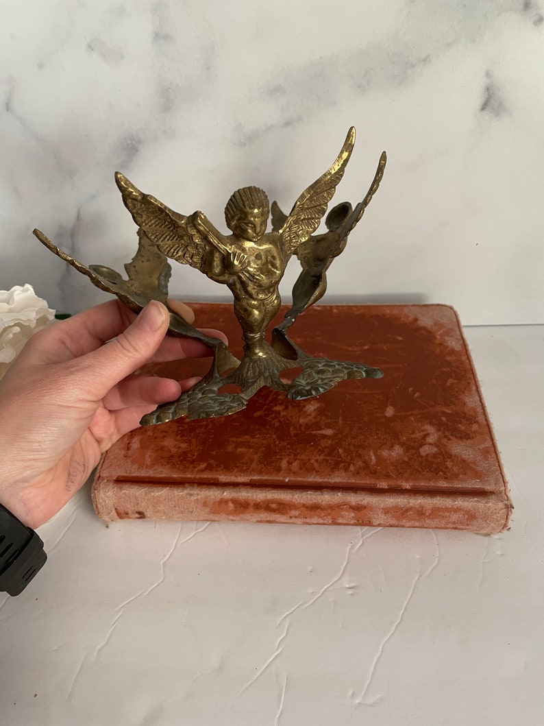 May include: A gold-toned metal angel figurine with wings outstretched. The angel is holding a musical instrument and is standing on a decorative base.