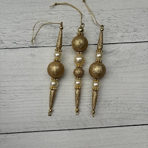 May include: Three gold Christmas ornaments with white pearl beads. Each ornament has a gold glitter finish and a loop for hanging.