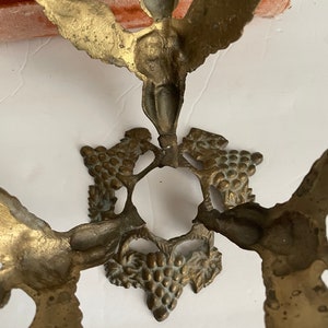 May include: A gold metal decorative stand with three winged cherubs and grapevine accents. The stand has a round top and a base with three legs.