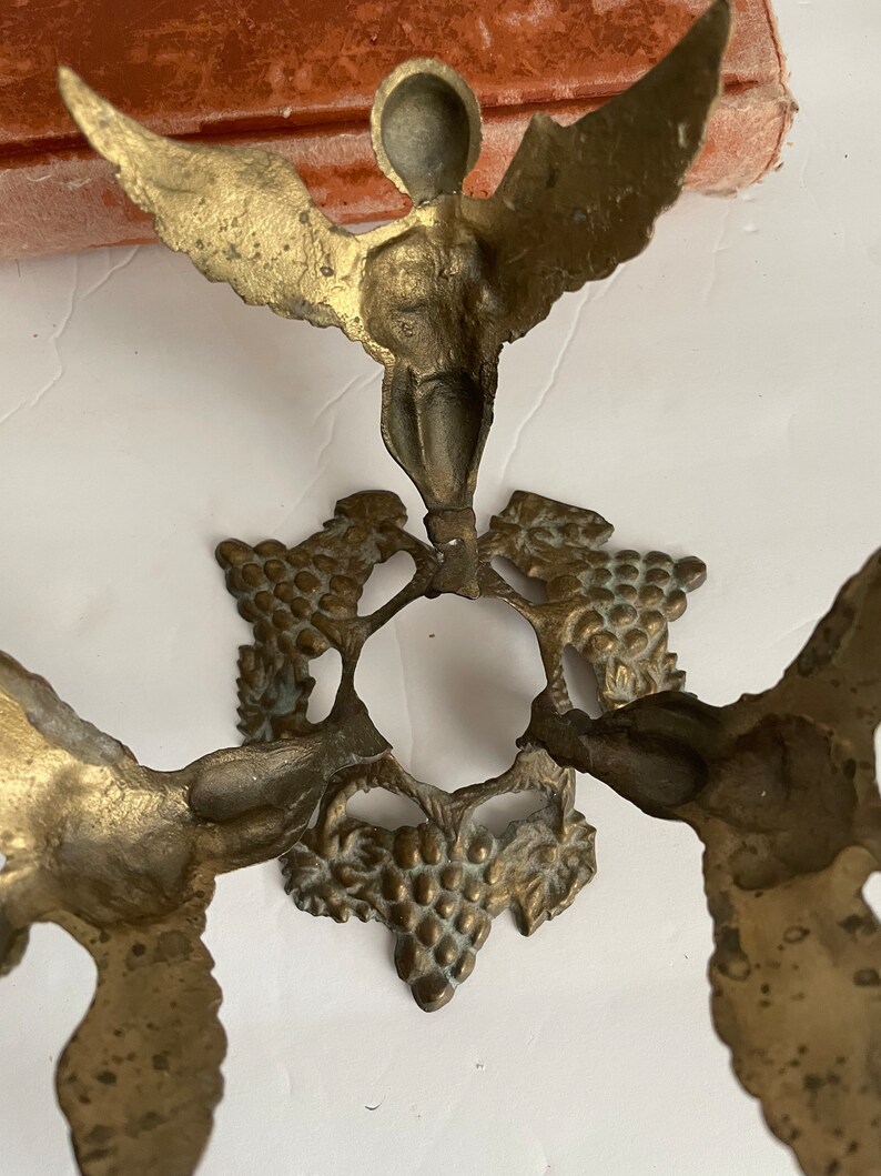 May include: A gold metal decorative stand with three winged cherubs and grapevine accents. The stand has a round top and a base with three legs.