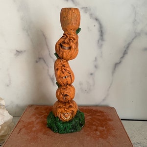 May include: A stacked pumpkin candle holder with four grinning faces. The candle holder is made of ceramic and has a green base.