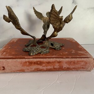 May include: A gold metal decorative stand with three angel figures. The stand has a round top and a base with a textured design.