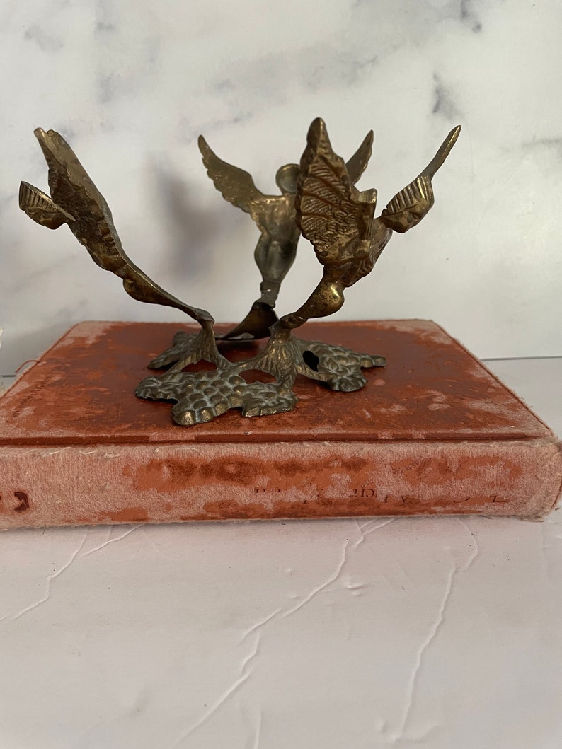 May include: A gold metal decorative stand with three angel figures. The stand has a round top and a base with a textured design.