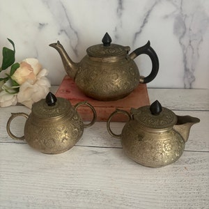 May include: A silver tea set with an ornate design. The teapot has a black handle and a black knob on the lid. The creamer and sugar bowl have black knobs on the lids.