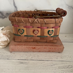 May include: A small, oval-shaped wicker basket with a wooden handle. The basket is decorated with pink and green ribbon and has three buttons with hearts on them.