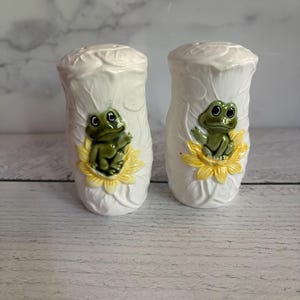May include: A pair of white ceramic salt and pepper shakers. Each shaker features a green frog perched on a yellow flower. The shakers have a textured leaf design on the top and sides.