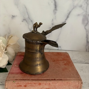 May include: A brass coffee pot with a camel finial on the lid. The pot has a spout and a handle. The pot is decorated with an intricate pattern. The pot is sitting on a pink book.