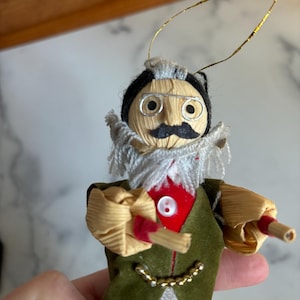 May include: A vintage corn husk doll ornament with a white beard, black hair, and a green jacket. The doll is wearing glasses and has a red button on its jacket. The doll is holding a pair of wooden sticks.