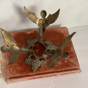 May include: A brass candle holder with three angel figures. The angels are holding up a circular base with a grapevine design.