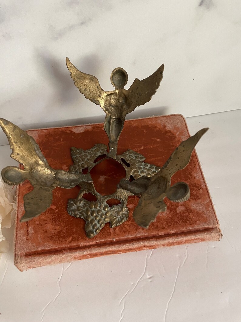 May include: A brass candle holder with three angel figures. The angels are holding up a circular base with a grapevine design.
