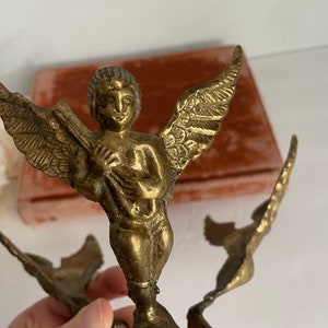 May include: A gold-toned metal sculpture of an angel with wings outstretched. The angel is holding a musical instrument and is standing on a base with a floral design.