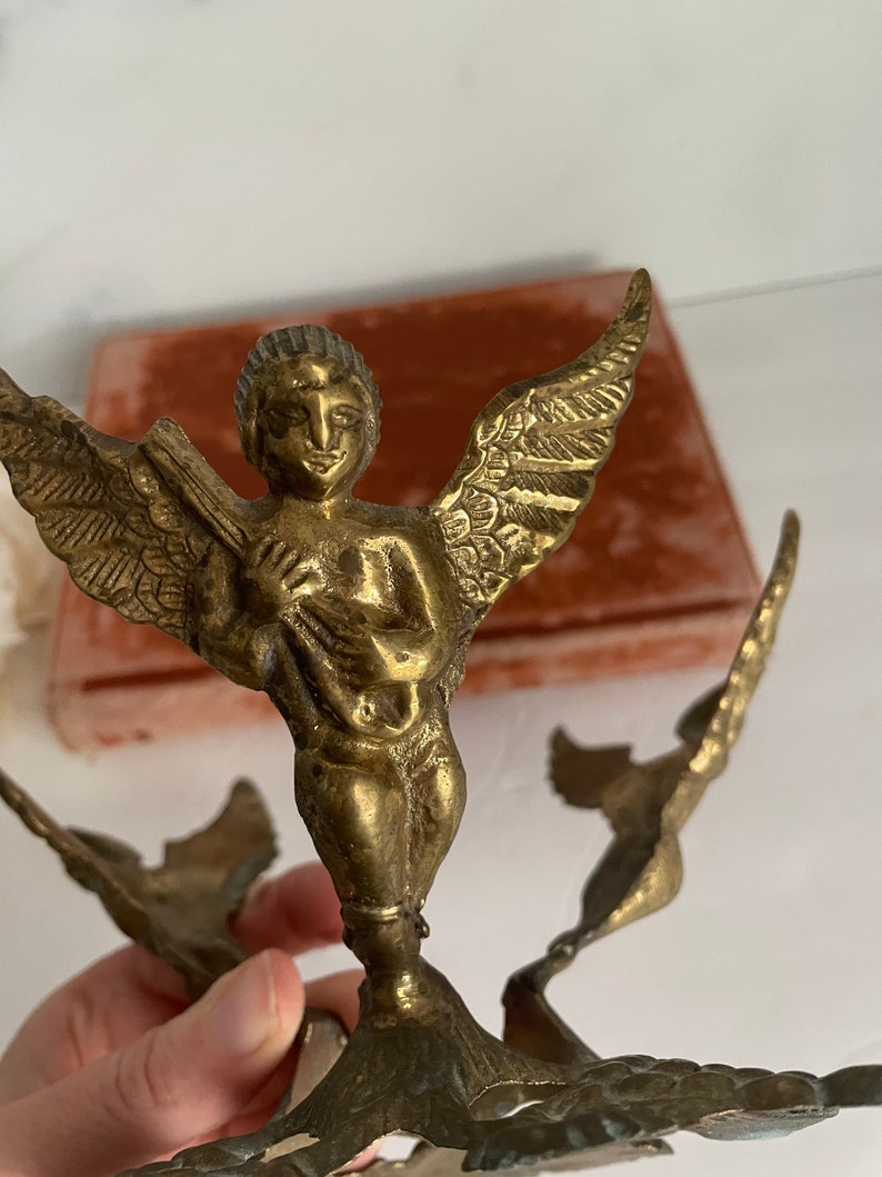 May include: A gold-toned metal sculpture of an angel with wings outstretched. The angel is holding a musical instrument and is standing on a base with a floral design.