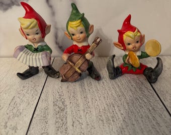 May include: Three ceramic elf figurines, each dressed in festive attire. One plays an accordion, another a violin, and the third a tambourine. They wear red and green outfits with pointed hats, and black shoes. The figurines are set against a light-coloured, wood-grain background.