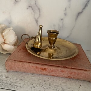 May include: A gold brass candlestick with a snuffer. The candlestick has a round base and a single candle holder. The snuffer is attached to the base with a hinge.