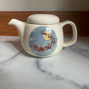 May include: White ceramic teapot with a lid, featuring a vintage illustration of Santa Claus flying in a sleigh pulled by reindeer against a blue and white snowy sky.