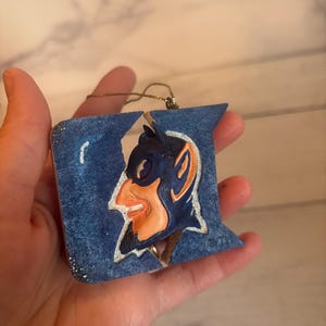 May include: Blue and orange Christmas ornament shaped like the Duke University logo with a cartoon devil's head. The ornament is made of felt and has a gold string for hanging.  The ornament is marked with a copyright symbol and TM.
