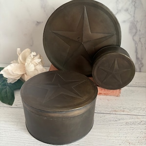May include: Three round metal containers with a star design. The containers are different sizes and are all a dark brown color.