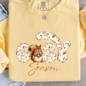 May include: A pale yellow crewneck sweatshirt featuring a graphic of a brown squirrel centered within the letters of the word COZY, with the word SEASON written in cursive below. The sweatshirt has a tag at the neck and long sleeves.