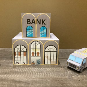 May include: A gray cardboard model of a bank building with a sign that reads "BANK" and a separate gray cardboard model of a bank truck with the words "BANK TRUCK" on the side.
