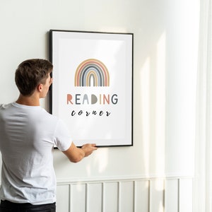 May include: A person hangs a framed print on a white wall. The print features a rainbow with the text "READING corner" in black lettering.